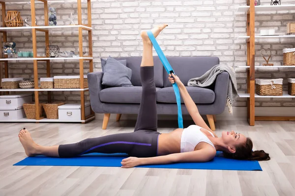 Young Woman Using Yoga Belt While Doing Exercise On Fitness Mat At Home