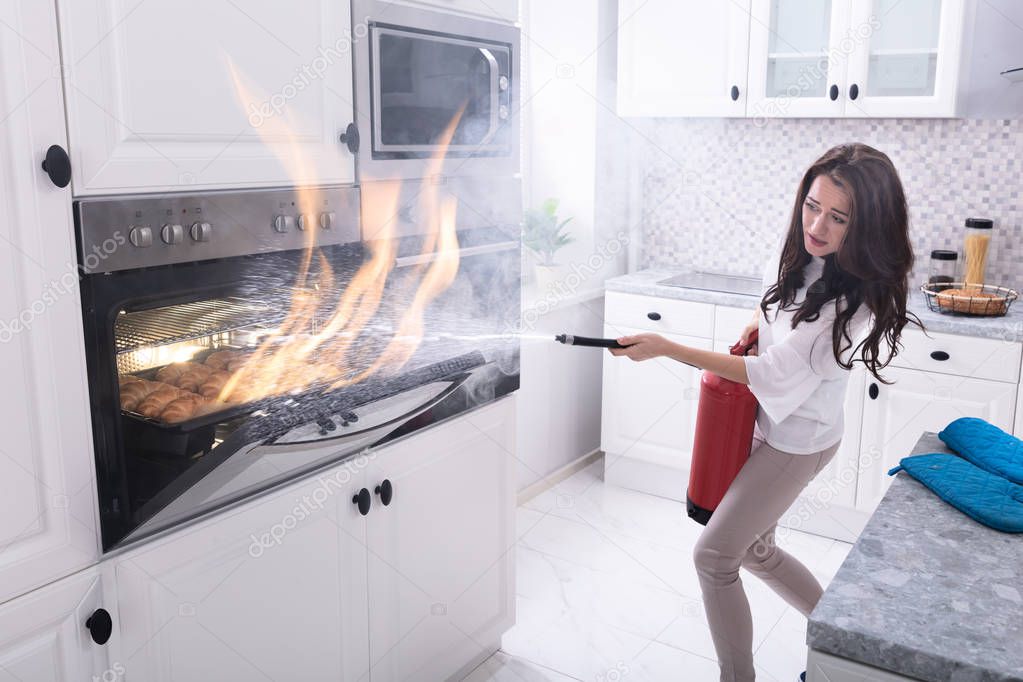 Woman Using Fire Extinguisher To Stop Fire Coming Out From Oven In Kitchen