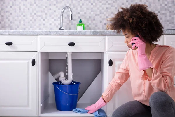 Young Woman Wearing Pink Gloves In Hand Calling Plumber In Front Of Leaking Sink Pipe