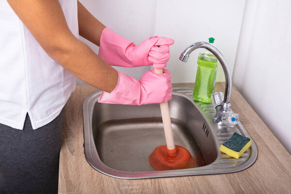 Close-up Of A Female Plumber Wearing Pink Gloves Using Plunger In The Kitchen Sink