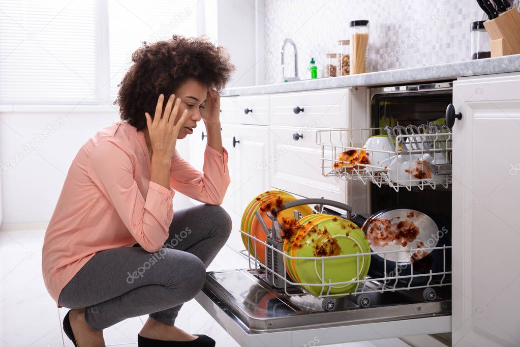 Worried Young Woman Looking At The Dirty Colorful Plates Arranged In The Dishwasher
