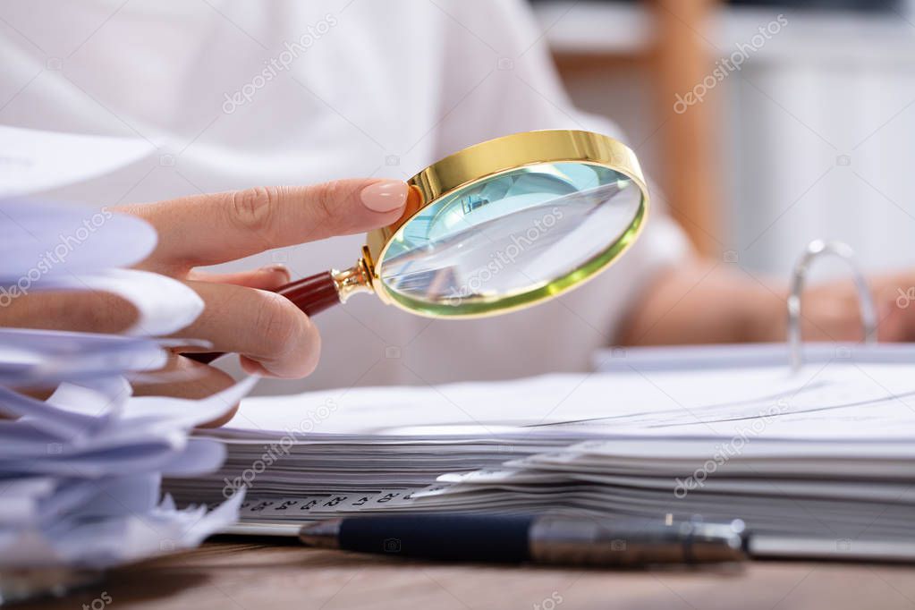 Close-up Of A Businesswoman's Hand Holding Magnifying Glass Over Invoice At Workplace