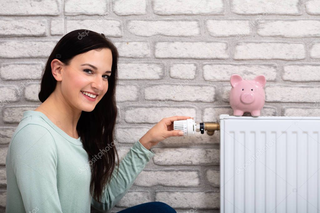 Young Smiling Woman Adjusting Thermostat With Piggy Bank On Radiator At Home