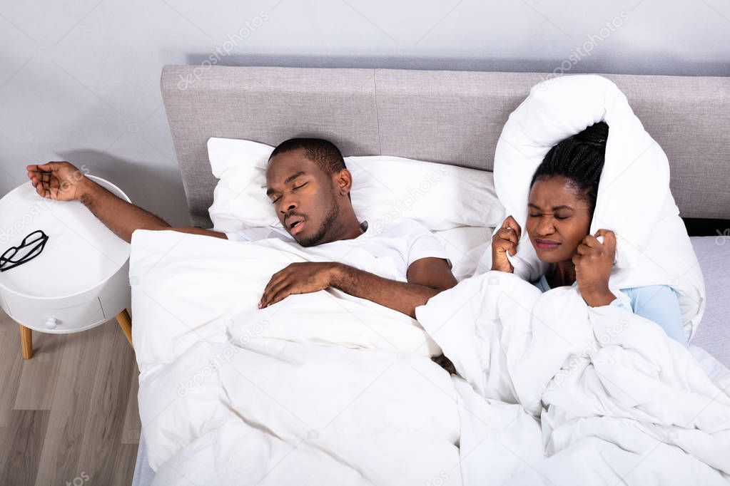 Disturbed African Woman Covering Her Ears With Pillow While Her Husband Snoring On Bed