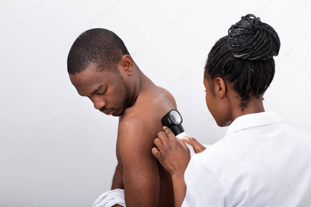 Young African Female Doctor Examining Pigmented Skin On Man's Back With Dermatoscope