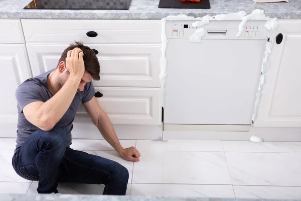 坐在受损洗碗机前的不安男子 泡沫来自它 — 图库照片
