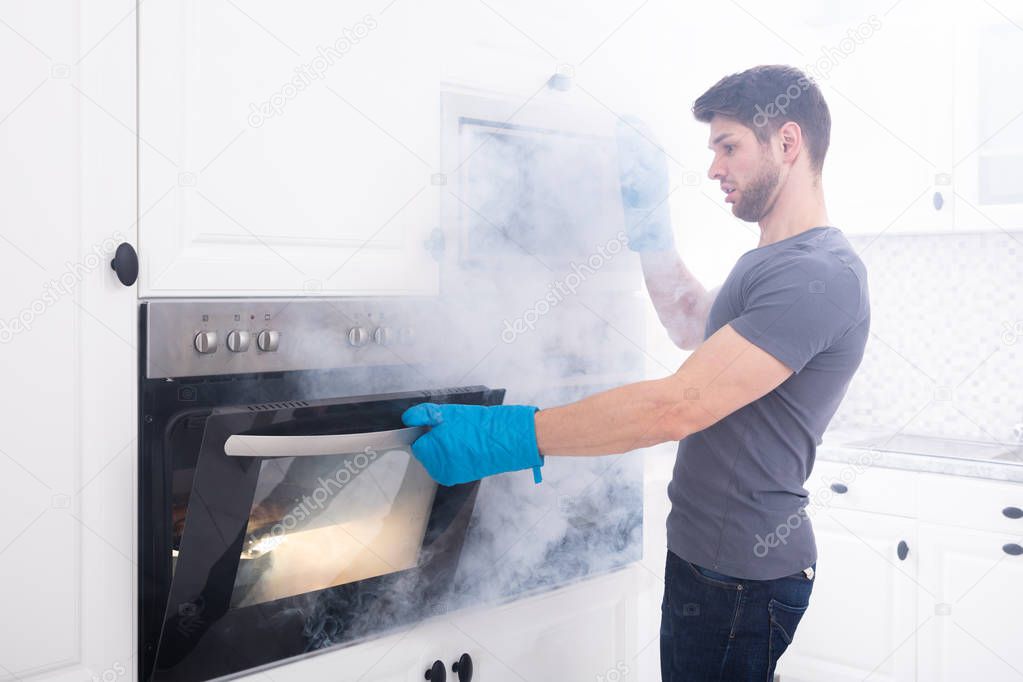 Young Man Wearing Gloves Opening Oven Filled With Smoke In Kitchen
