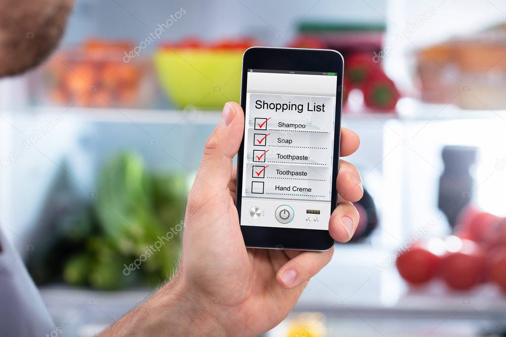 Close-up Of Human Hand Checking Shopping List On Mobilephone In Front Of Refrigerator