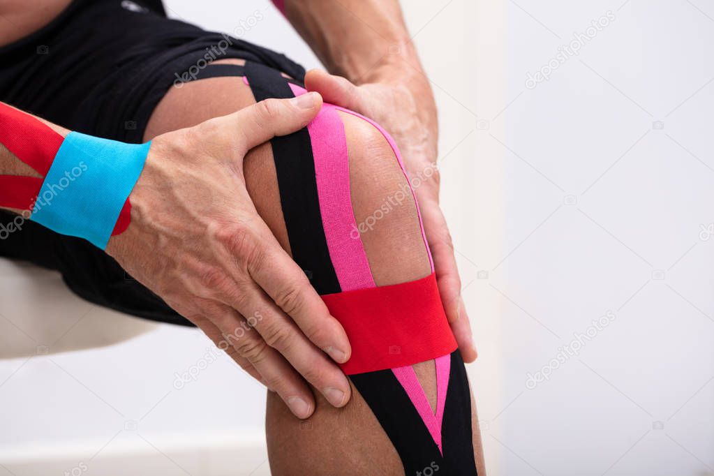 Man Sitting On Table Applying Kinesiology Tape On His Knee