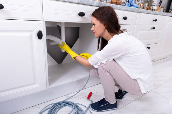 Side View Of A Young Woman Cleaning Clogged Sink Pipe In Kitchen