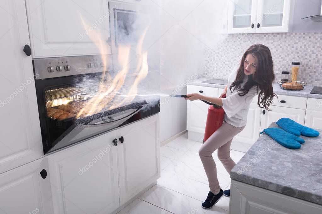 Woman Using Fire Extinguisher To Stop Fire Coming Out From Oven In Kitchen