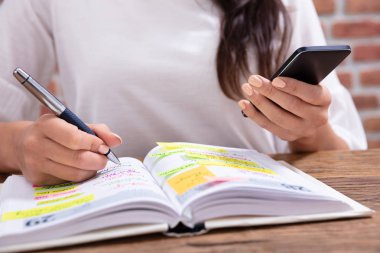 Close-up Of A Businesswoman Holding Cellphone Writing Schedule In The Diary With Pen clipart