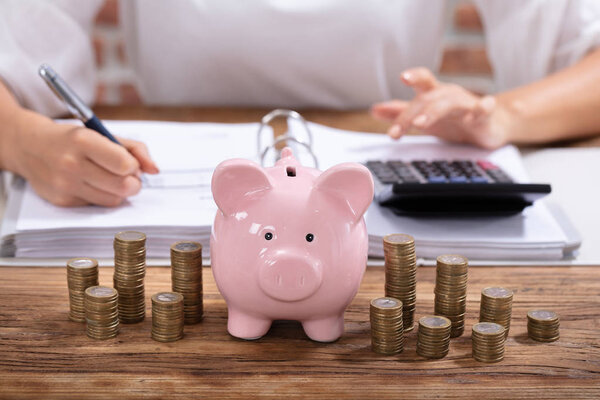 Stacked Coins And Pink Piggy Bank In Front Of Businesswoman Calculating Invoice On Wooden Desk