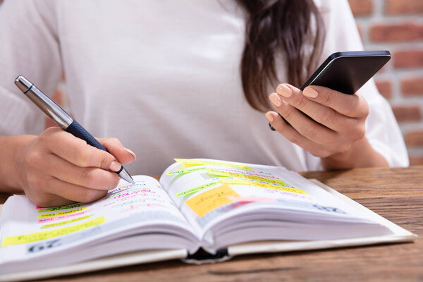 Close-up Of A Businesswoman Holding Cellphone Writing Schedule In The Diary With Pen