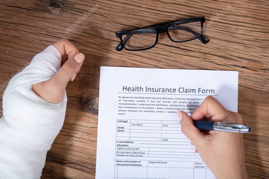 Close-up Of Businesswoman With White Bandage Hand Filling Health Insurance Claim Form On Wooden Desk