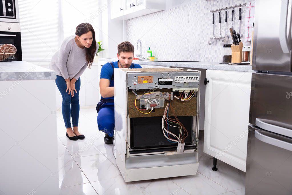 Smiling Woman Behind Technician Repairing Dishwasher In Kitchen