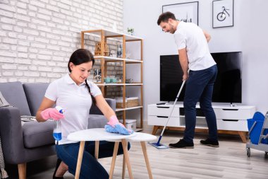 Gülümseyen iki genç işçi oturma odasındaki masaları temizleyip yerleri paspaslıyor.