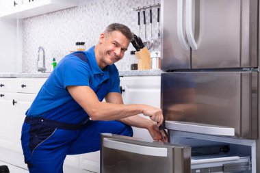 Mature Male Serviceman Repairing Refrigerator With Toolbox In  Kitchen clipart
