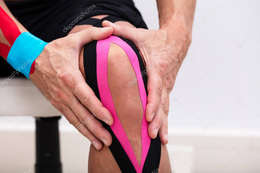 Man Sitting On Table Applying Kinesiology Tape On His Knee