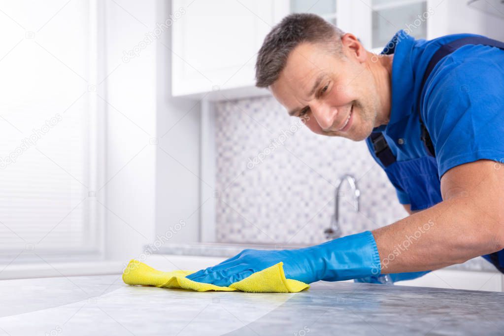 Mid Section Of A Male Janitor Cleaning Dirty Kitchen Counter With Spray Bottle And Napkin