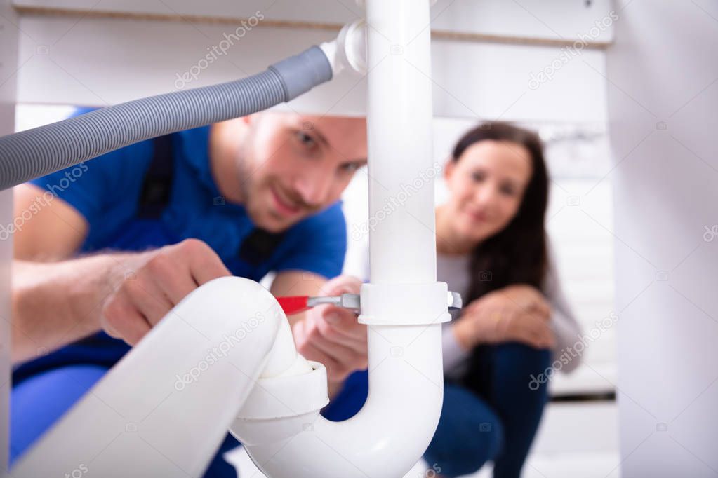 Young Woman Looking At Male Plumber Fixing Sink Pipe In Kitchen