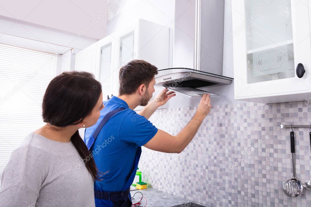 Pretty Young Woman Looking At Repairman Fixing Kitchen Extractor Filter Over Kitchen Counter