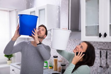 Worried Young Woman Calling Plumber On Cellphone While Man Collecting Leakage Water From Ceiling clipart