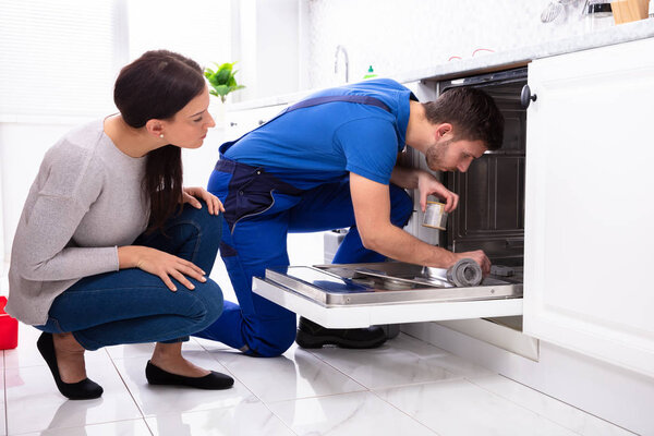 Young Woman Looking At Repairman Repairing Dishwasher In Kitchen