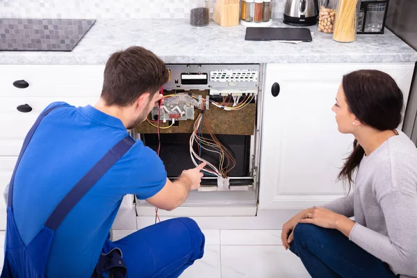 キッチンでデジタルマルチメーターで食器洗い機をチェックする男性技術者を見て女性 — ストック写真