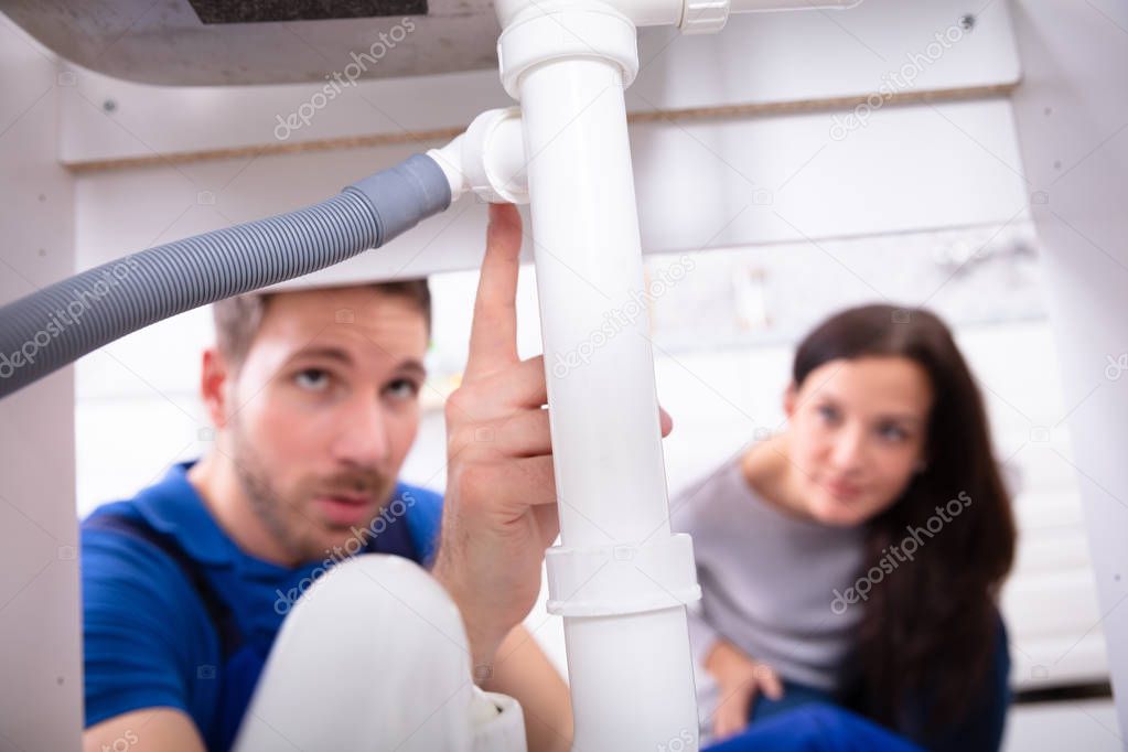 Young Male Plumber Showing Damage In Sink Pipe To Woman At Home