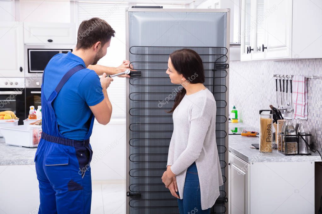 Woman Looking At Technician Repairing Refrigerator With Screwdriver In Kitchen