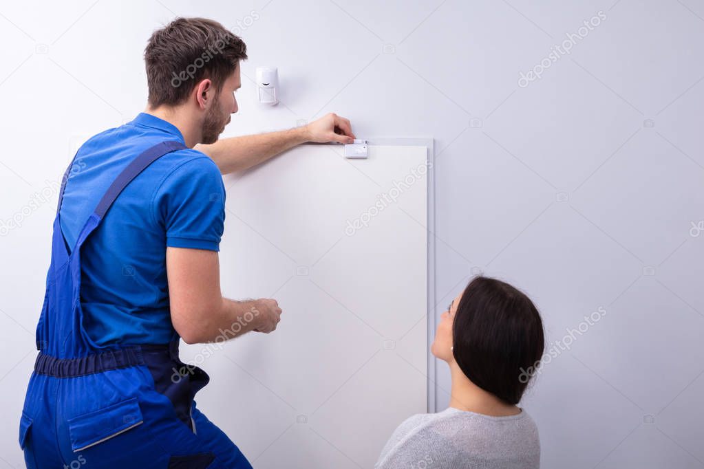 Happy Woman Looking At Electrician Installing Security System Door Sensor On Wall
