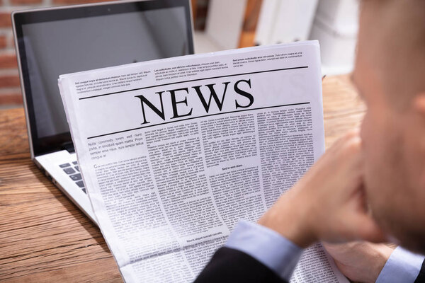 Close-up Of A Man's Hand Holding Newspaper Over Wooden Desk