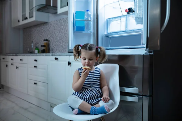 Menina Bonito Comendo Bolo Enquanto Sentado Frente Geladeira Aberta Cozinha — Fotografia de Stock