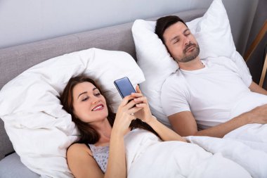 Onu kocası yatak odasında yatakta Sleeping Iken Smartphone kullanarak gülümseyen kadın