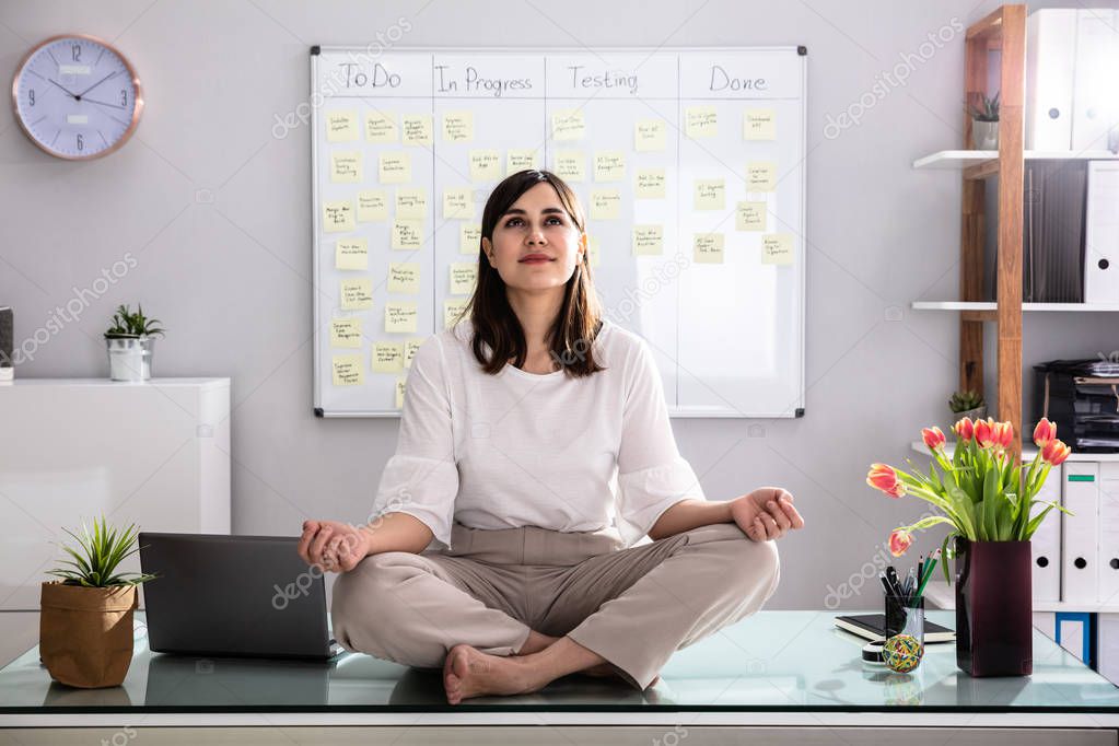 Young Businesswoman Sitting On Desk Meditating In Office