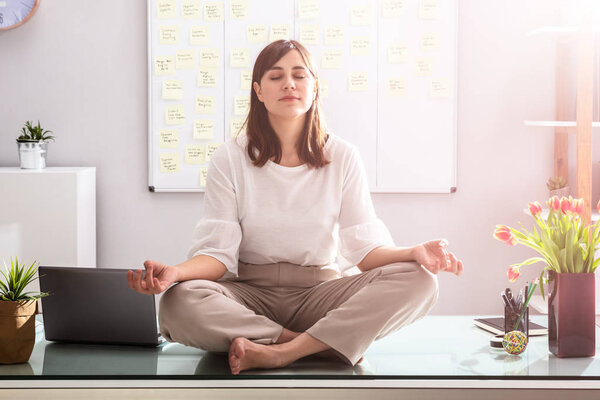 Young Businesswoman Sitting On Desk Meditating In Office