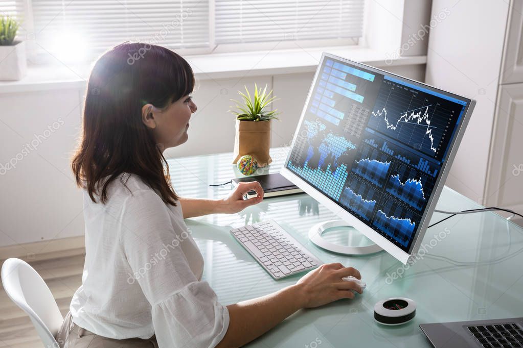 Young Female Stock Market Broker Analyzing Graph On Computer At Workplace