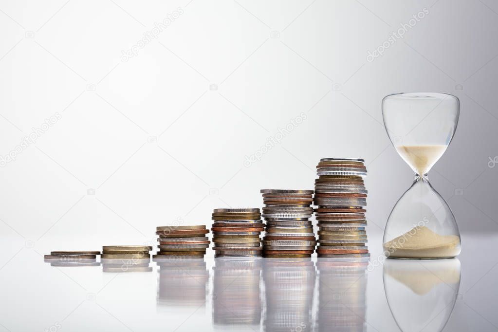 Close-up Of Hourglass Near Stack Of Coins Over Desk