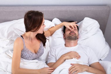 Disturbed Young Woman Holding Her Husband's Nose To Stop Him From Snoring On Bed clipart