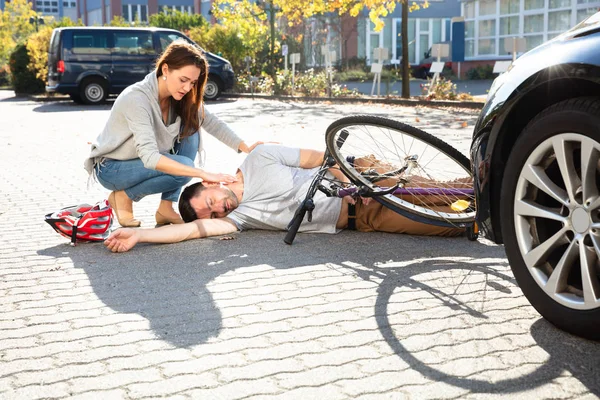 年轻女子看着无意识的男子自行车运动员躺在汽车附近的大街上 — 图库照片