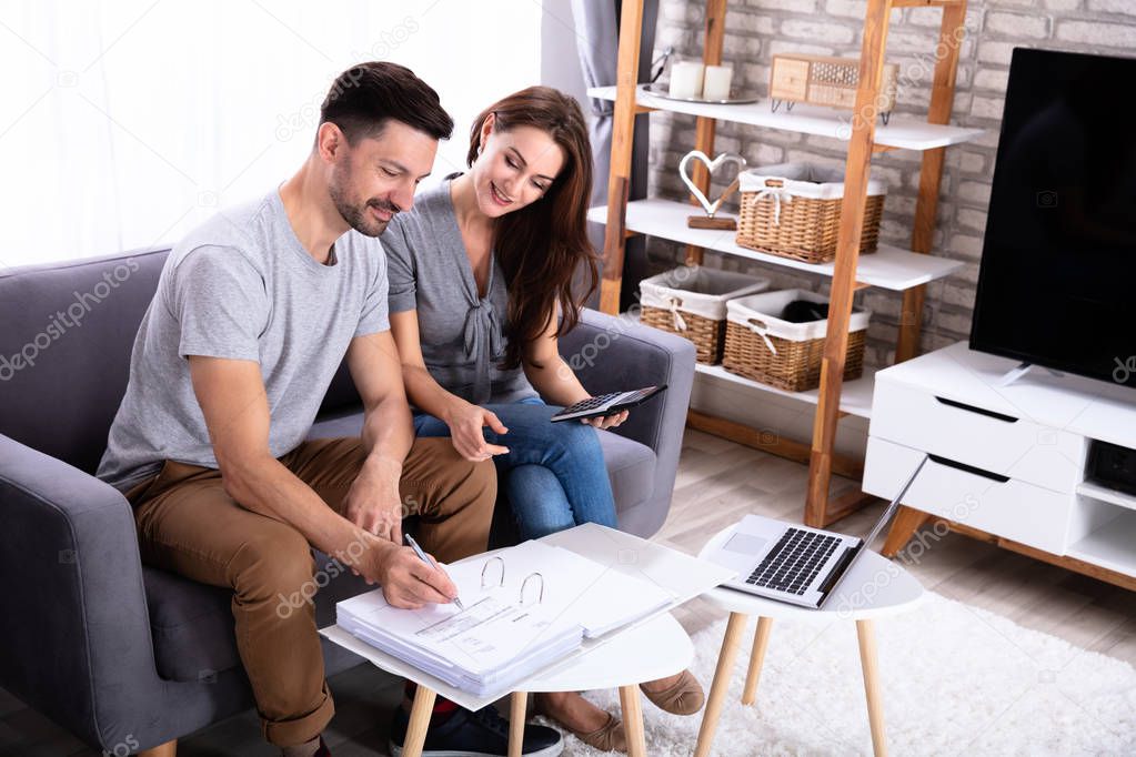 Smiling Young Couple Sitting On Sofa Calculating Invoice At Home
