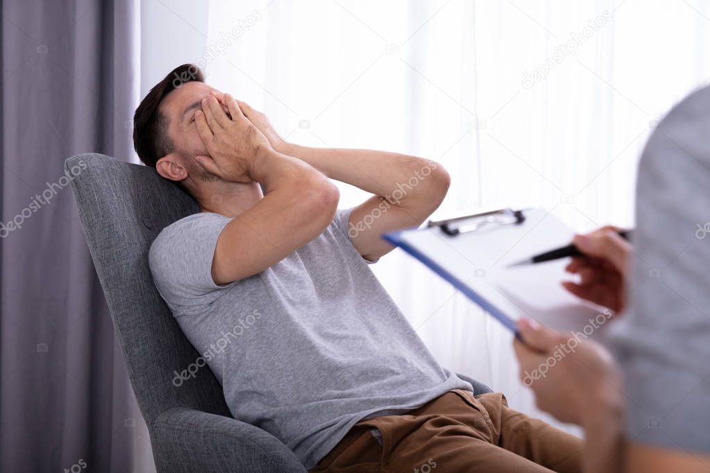 Female Psychologist With Clipboard Sitting Near Young Man Suffering From Depression