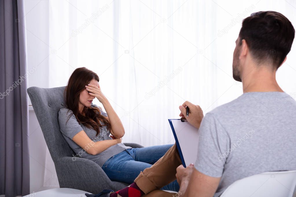 Male Psychologist With Clipboard Sitting Near Young Woman Suffering From Depression