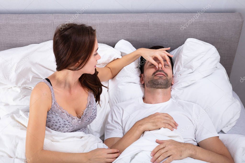 Disturbed Young Woman Holding Her Husband's Nose To Stop Him From Snoring On Bed