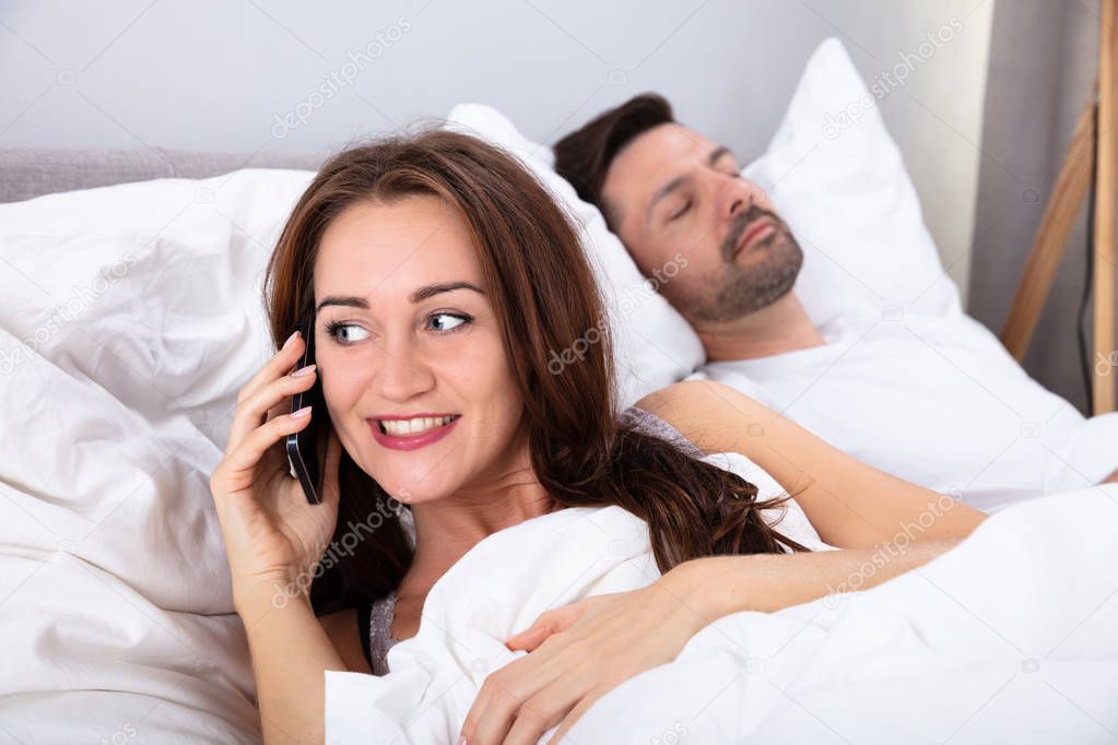 Smiling Young Woman Talking On Smartphone Besides Her Husband Sleeping On Bed In Bedroom