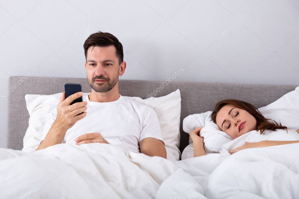 Smiling Man Using Smartphone While His Wife Sleeping On Bed In Bedroom