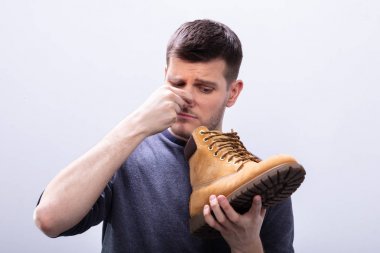 Close-up Of A Man Covering His Nose While Holding Stinky Shoe Against White Background clipart