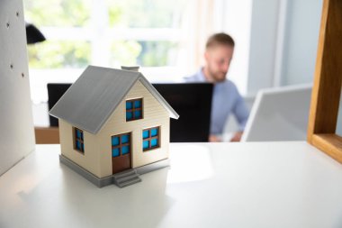 House Model On Shelf And Man Working In Office At Background clipart