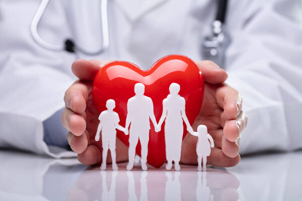 Close-up Of A Doctor's Hand Protecting White Family Paper Cut Out With Heart Shape
